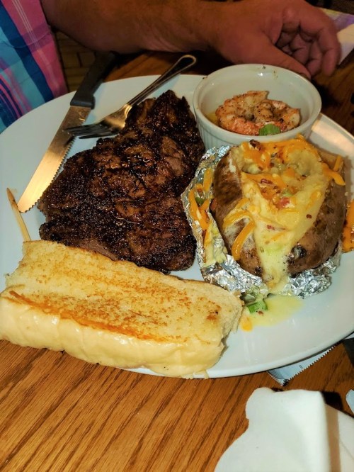 A plate with a grilled steak, baked potato topped with cheese, shrimp, and a slice of toasted bread.