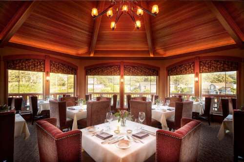 Elegant restaurant interior with wooden beams, large windows, and tables set for dining. Warm lighting creates a cozy atmosphere.