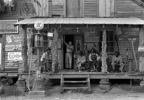 A vintage gas station with people sitting on the porch, surrounded by old advertisements and wooden structures.