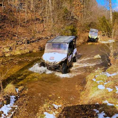 Two off-road vehicles drive through a shallow stream in a wooded area, with snow patches on the ground.