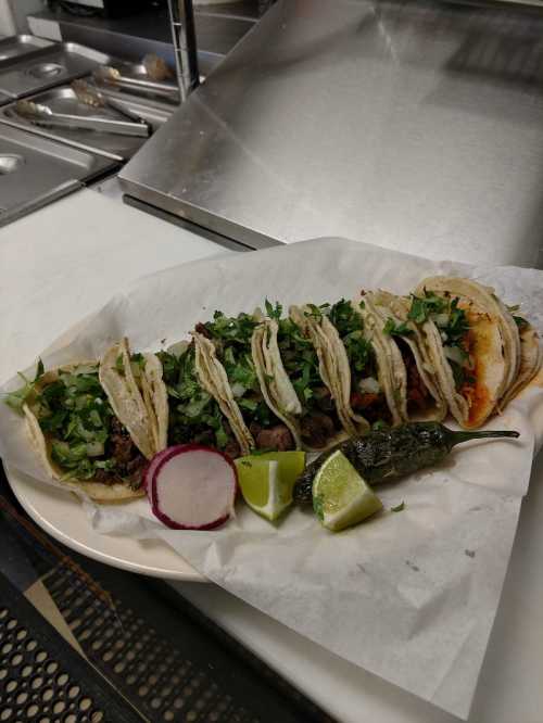 A plate of tacos topped with cilantro, served with radish, lime, and a jalapeño pepper on a white paper-lined dish.