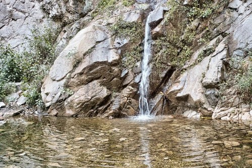 A small waterfall cascades down rocky terrain into a clear pool, surrounded by lush greenery.