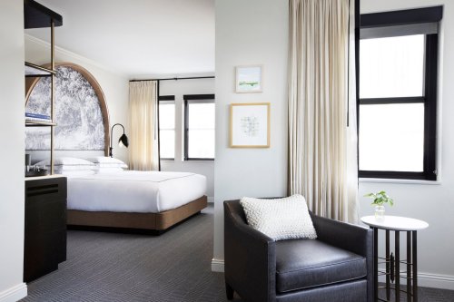Modern hotel room featuring a bed, armchair, and large windows with natural light. Elegant decor and neutral colors.