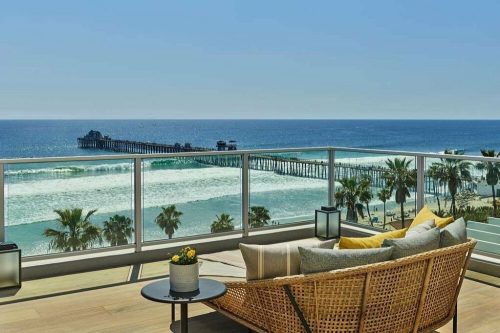 A coastal view from a balcony featuring a wicker chair, ocean waves, palm trees, and a pier in the distance.