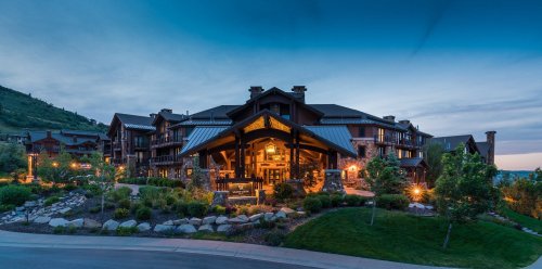 A large, rustic lodge surrounded by greenery, illuminated at dusk with a clear blue sky above.