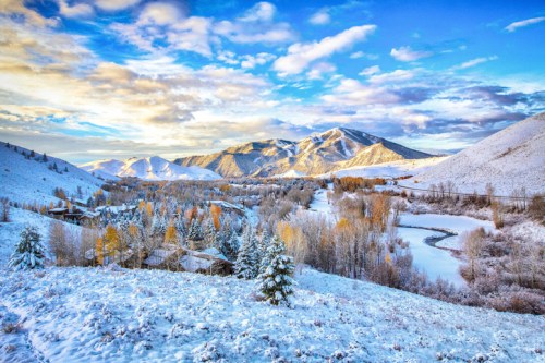 A snowy landscape with mountains, a river, and colorful trees under a bright blue sky with fluffy clouds.