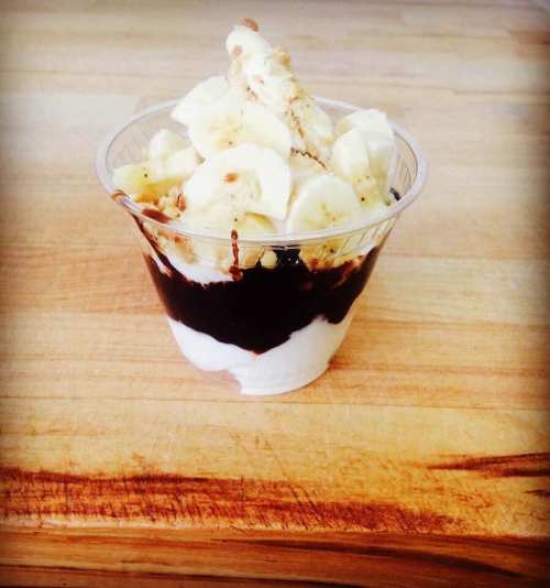A clear cup filled with layers of yogurt, banana slices, and chocolate sauce, placed on a wooden surface.