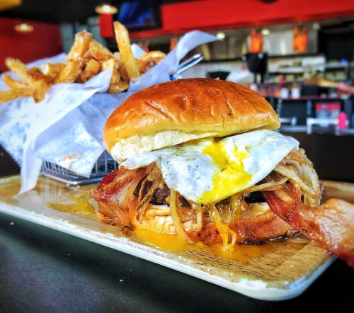 A juicy burger topped with a fried egg and bacon, served with a side of crispy fries on a wooden platter.