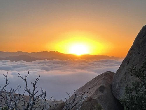 A stunning sunset over mountains, with the sun glowing above a sea of clouds and rocky foreground.