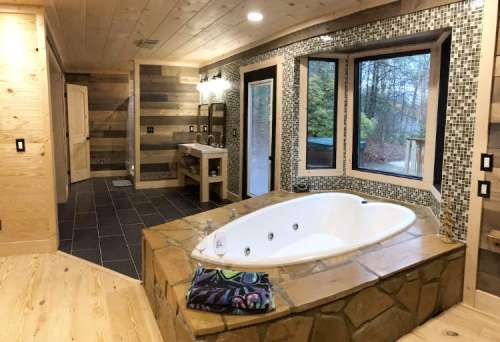 A modern bathroom featuring a large jetted tub, stone accents, wooden walls, and a window with a view.