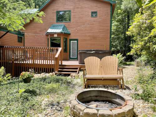 A wooden house surrounded by trees, featuring a deck, hot tub, and a fire pit with two Adirondack chairs nearby.