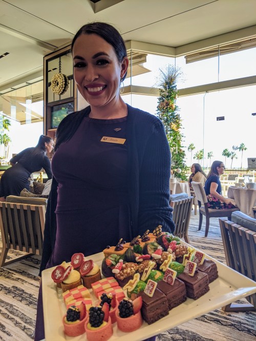 A smiling server holds a tray of colorful desserts at a festive event, with a decorated tree in the background.