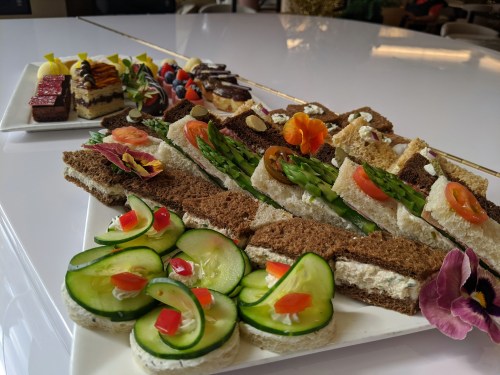 A platter of assorted sandwiches with vegetables, garnished with flowers, alongside cucumber slices and colorful desserts.