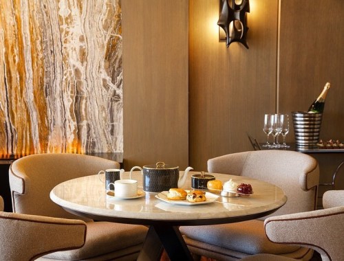 A stylish table set with a teapot, cups, pastries, and a dessert, surrounded by elegant chairs in a warm-toned room.
