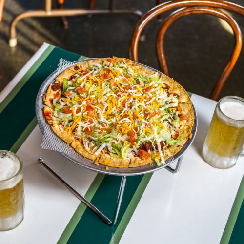 A colorful taco pizza topped with lettuce, tomatoes, cheese, and served with two glasses of beer on a table.