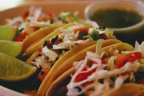 A close-up of three tacos filled with cabbage, tomatoes, and cilantro, served with lime wedges and a side of green salsa.