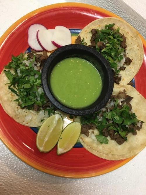 Three tacos filled with meat, topped with onions and cilantro, served with green salsa, lime wedges, and radish slices.