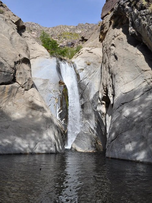 A serene waterfall cascades into a clear pool, surrounded by rocky cliffs and sparse greenery under a blue sky.