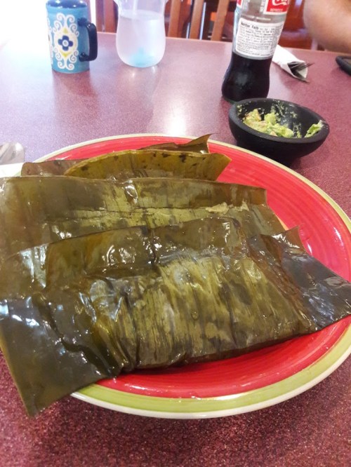 A plate with several green banana leaves, accompanied by a small bowl of guacamole and a drink in the background.