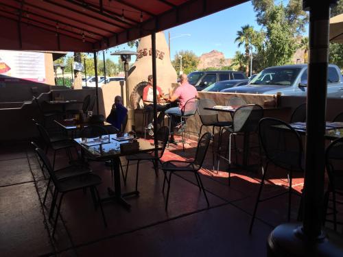 Outdoor dining area with empty tables, a man sitting near a fireplace, and parked cars in the background.