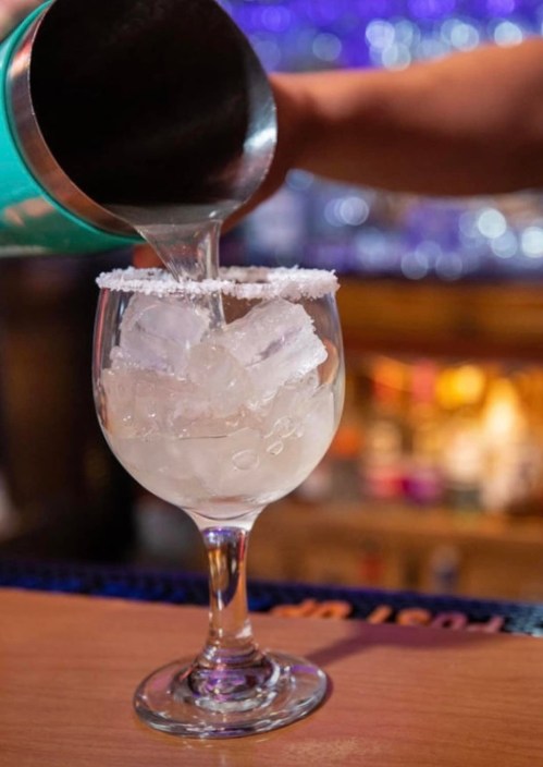 A cocktail is being poured into a glass with ice and a salted rim, set against a blurred bar background.