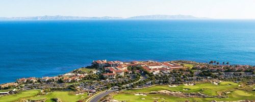 Aerial view of a coastal resort with golf courses, overlooking the ocean and distant islands under a clear blue sky.