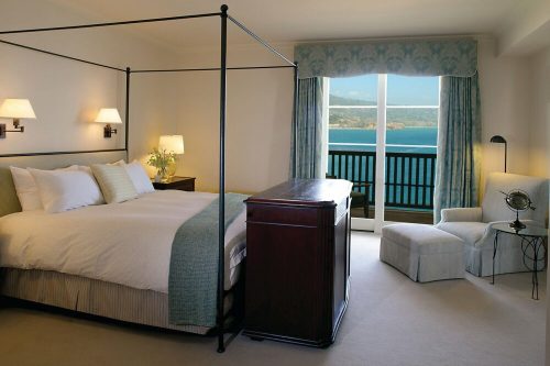 A stylish bedroom with a four-poster bed, a wooden dresser, and a view of the ocean through large windows.