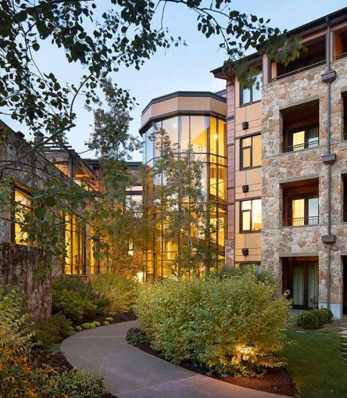 A modern building with large glass windows surrounded by lush greenery and a winding pathway at dusk.