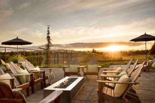 A serene outdoor patio with wooden chairs, a fire pit, and umbrellas, overlooking a sunset and scenic landscape.