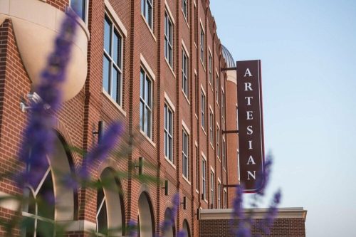 A close-up of a brick building with a sign reading "Artesian," framed by purple flowers in the foreground.