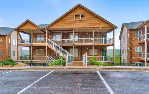 A wooden multi-story building with balconies, surrounded by greenery and a parking area in front.