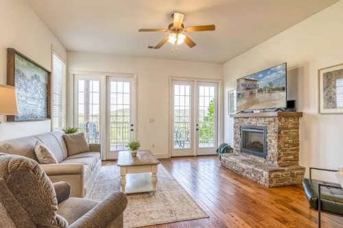 Cozy living room with a stone fireplace, TV, and large windows overlooking a scenic view. Light-colored furniture and wood floors.