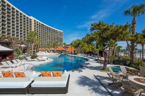 A sunny resort pool area with lounge chairs, palm trees, and a modern building in the background.