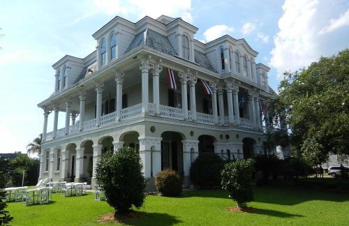A large, historic white mansion with multiple columns, flags, and a lush green lawn under a blue sky.