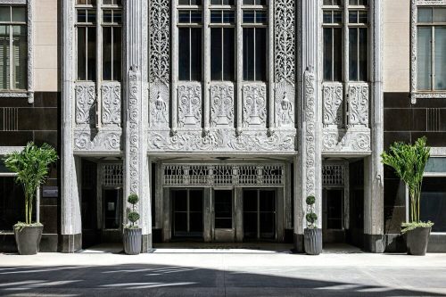Art Deco building entrance with intricate carvings and potted plants on either side, set in a sunny urban environment.