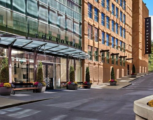 Entrance of The Fontaine hotel, featuring a glass canopy, planters with flowers, and a modern architectural design.