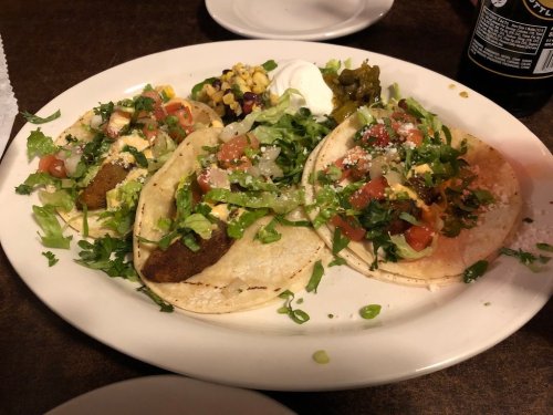 A plate of three tacos topped with lettuce, tomatoes, cheese, and cilantro, served with sides of salsa and sour cream.