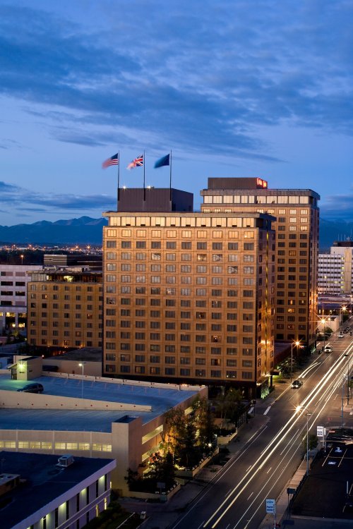 A cityscape at dusk featuring a tall hotel with flags, surrounded by buildings and a busy street with light trails.