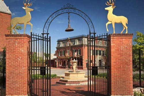 A decorative gate with golden deer statues leads to a brick building and fountain in a sunny outdoor setting.