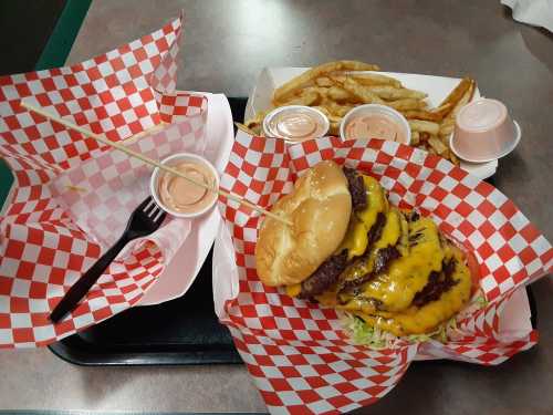 A large cheeseburger with multiple patties on a tray, accompanied by fries and dipping sauces in red-checkered wrappers.