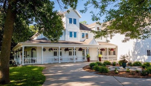 A charming white house with a porch, surrounded by greenery and flower pots, set on a sunny day.