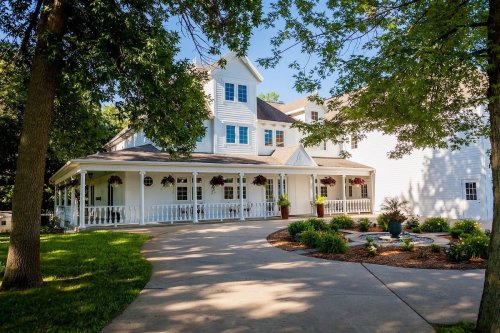 A charming white house with a porch, surrounded by greenery and flower pots, set on a sunny day.