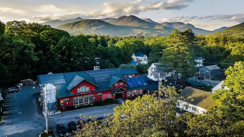 Aerial view of a red building with solar panels, surrounded by trees and mountains under a partly cloudy sky.