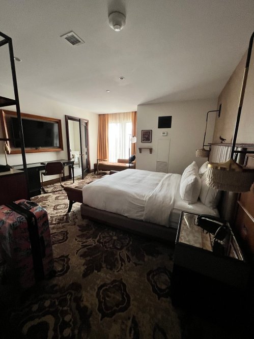 A modern hotel room featuring a bed, TV, desk, and large window with natural light. Stylish decor and furnishings.