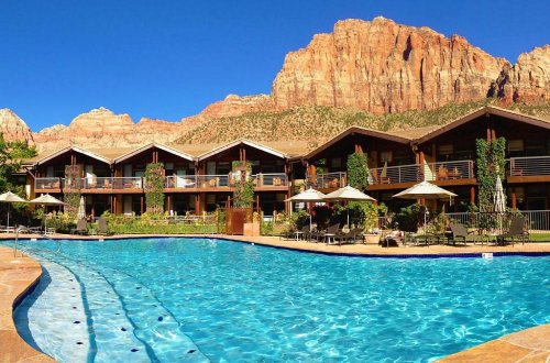 A scenic hotel with a pool, surrounded by mountains and clear blue skies. Sun loungers and umbrellas are by the water.