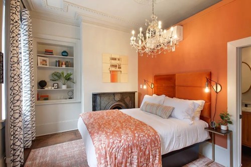 Cozy bedroom with orange accent wall, chandelier, and stylish decor, featuring a plush bed and bookshelf.