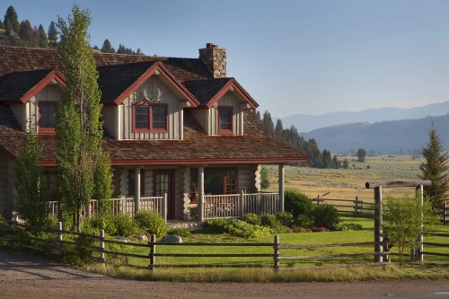 A charming log cabin with red accents, surrounded by greenery and mountains in the background under a clear blue sky.