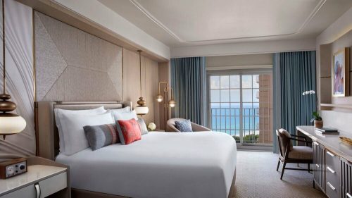 A modern hotel room with a large bed, ocean view, and stylish decor, featuring a desk and comfortable seating area.