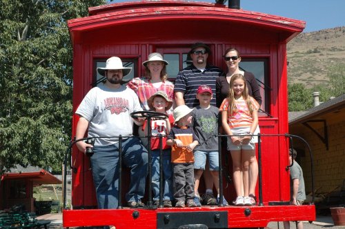 A group of seven people, including children and adults, pose together on a red train car in a sunny outdoor setting.