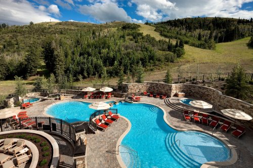 A scenic outdoor pool area with lounge chairs, surrounded by lush greenery and hills under a partly cloudy sky.